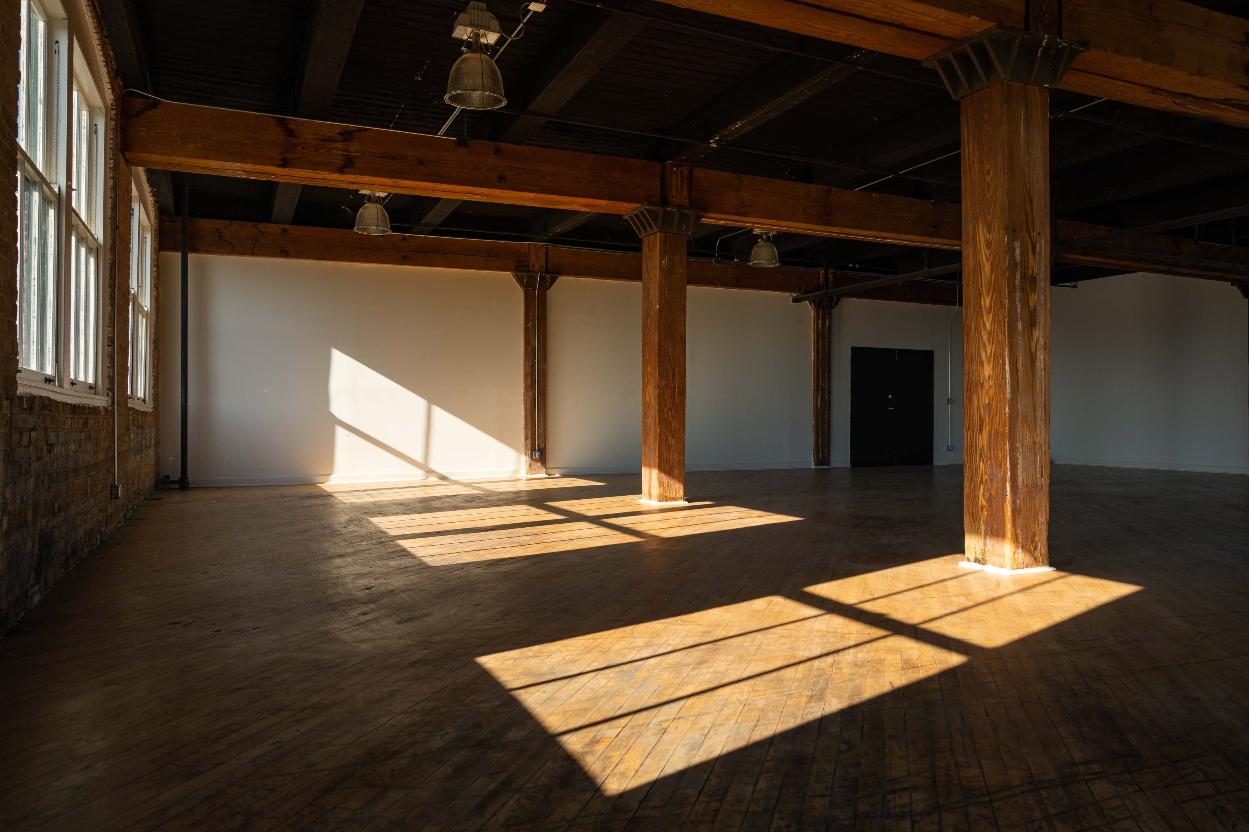 A dancer poses with one leg extended to the side in a spacious, well-lit studio with wooden floors and exposed brick walls.