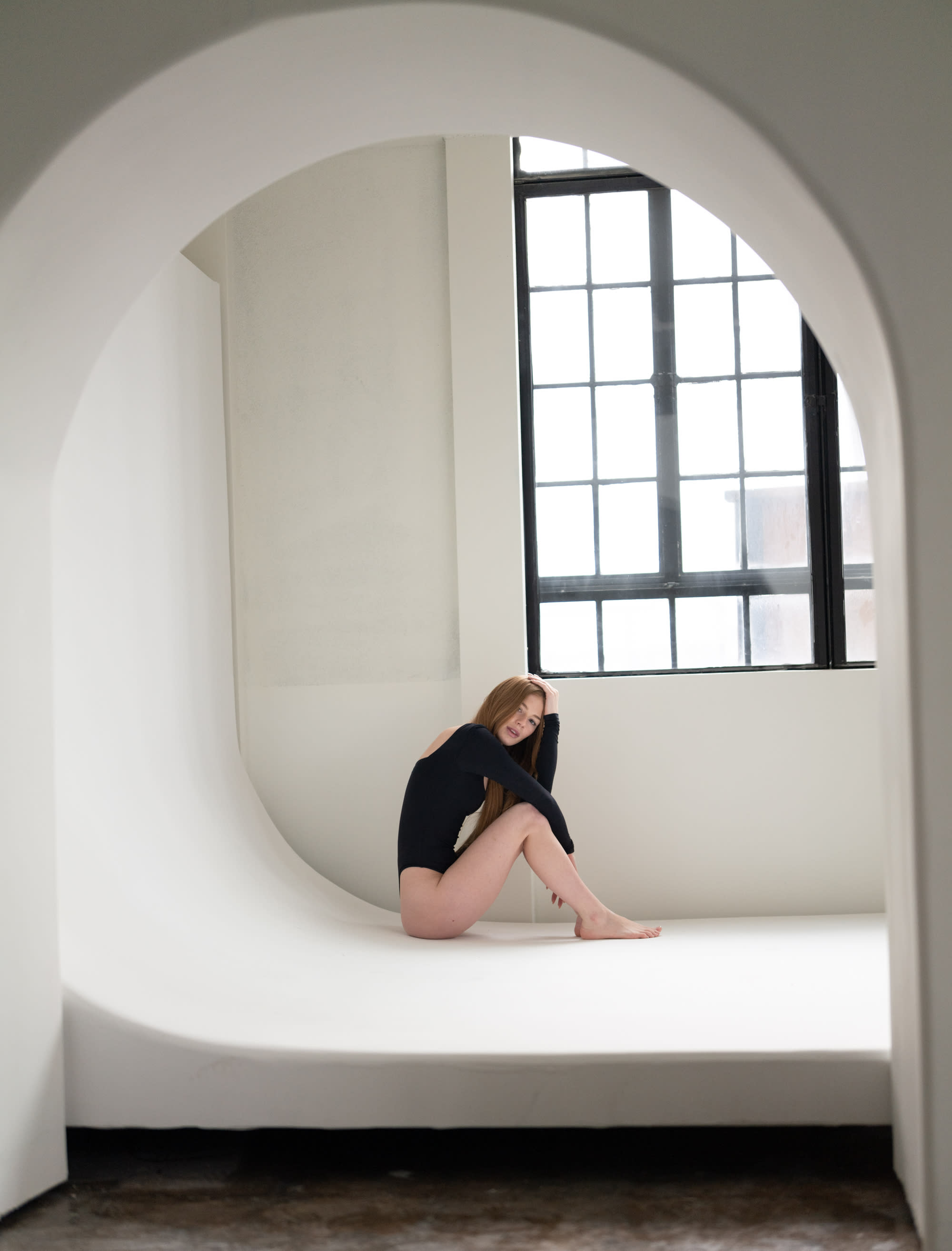 The image shows a minimalist studio space with a curved white backdrop and large windows letting in natural light.