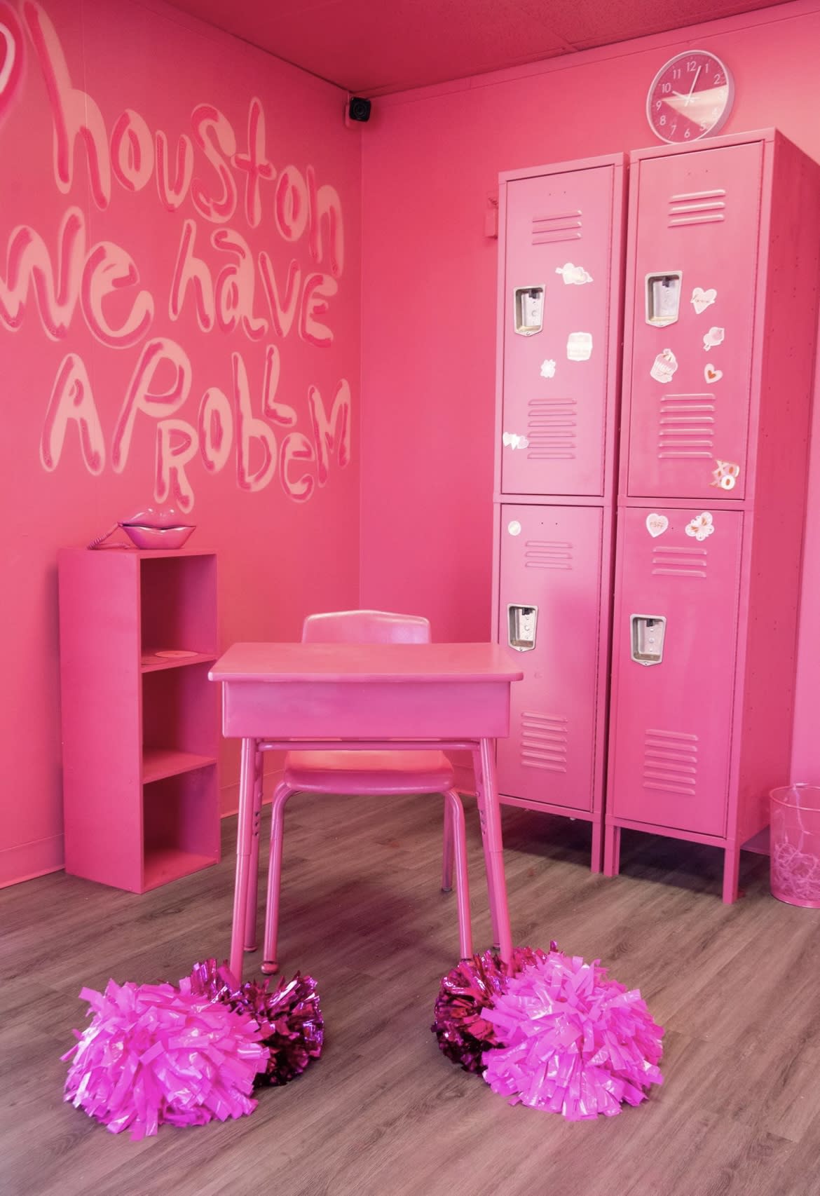 A person sits at a pink desk in a room adorned with pink lockers and decor, holding a vintage phone during a photoshoot.