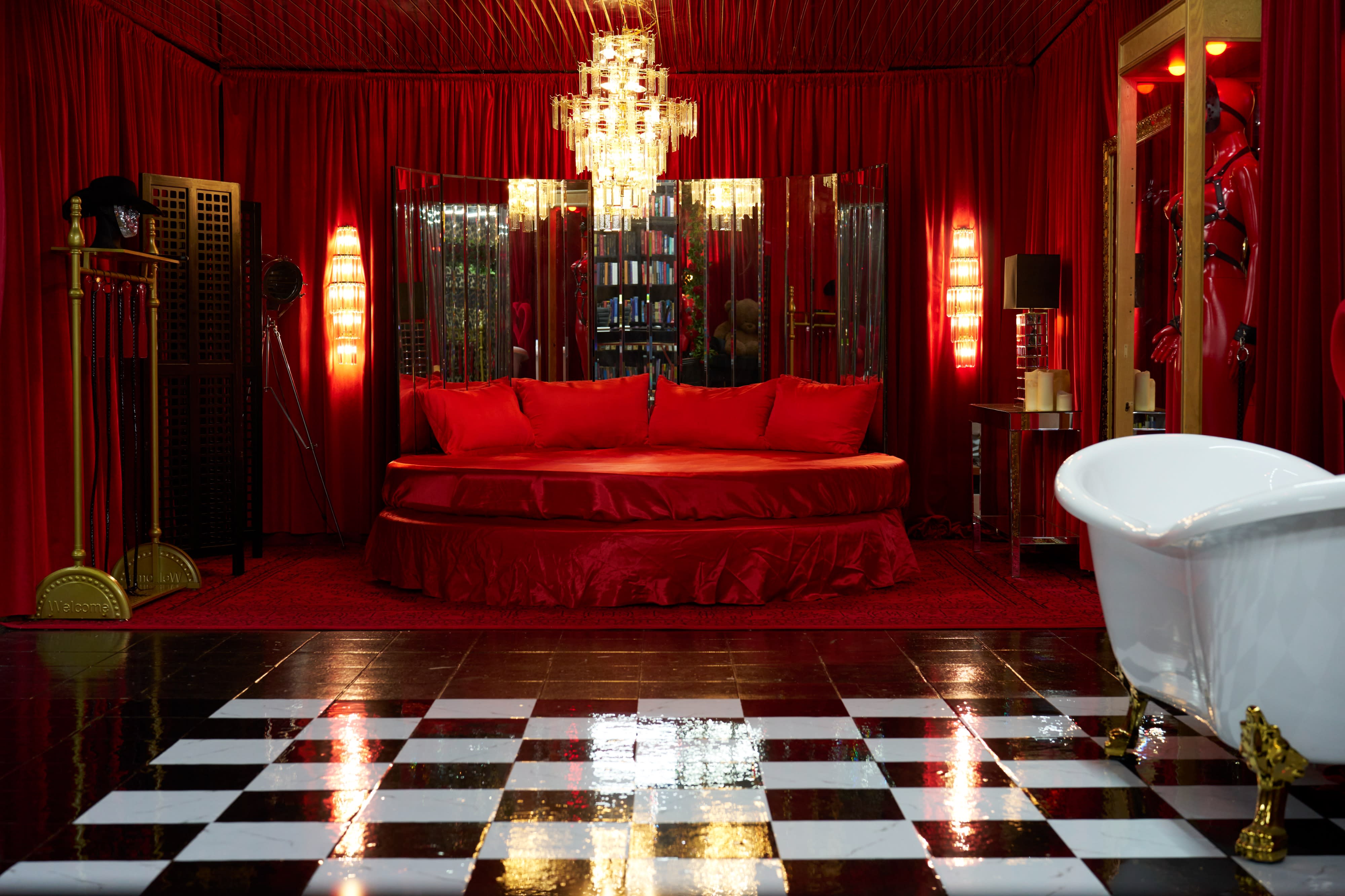 A person lies on a red bed with a floral arrangement in their arms against a dark background.