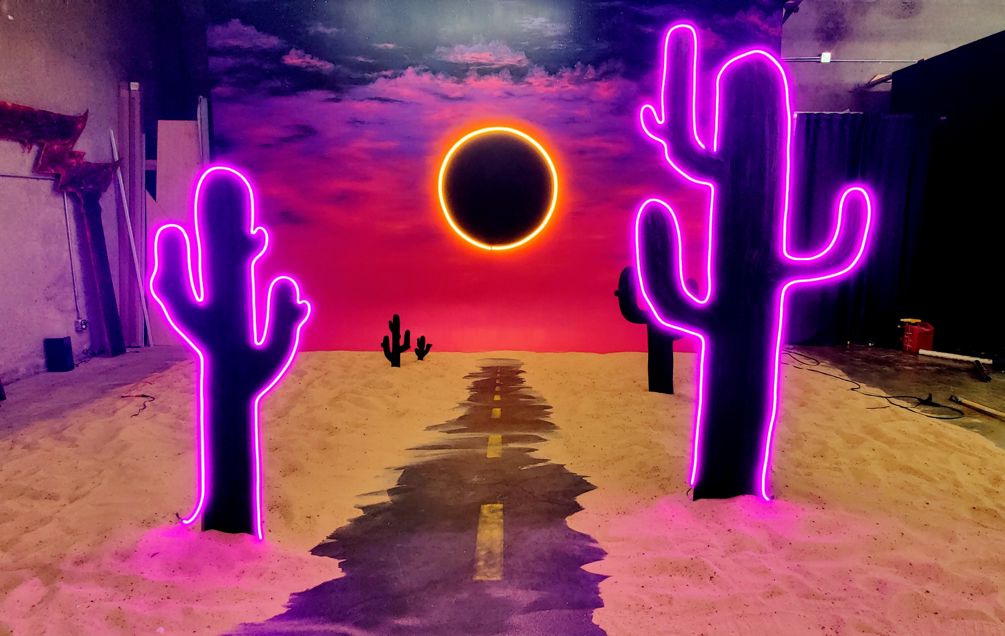 A video shoot features a man standing confidently in the center, flanked by two women in cowboy attire, set against neon cactus backdrops in a sandy environment.