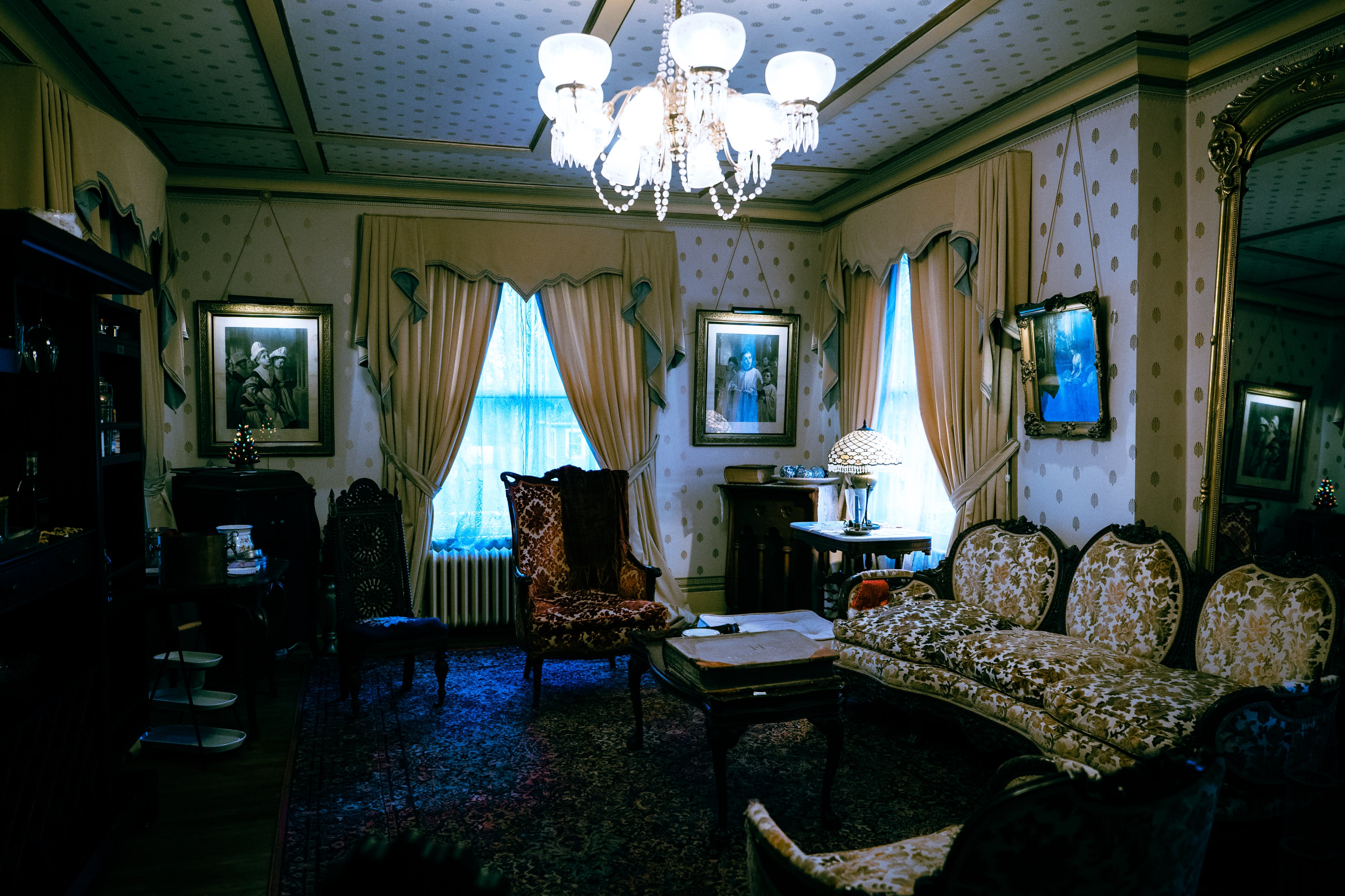 A woman in a lace dress sits by a window, gently pulling back a curtain while gazing outside.