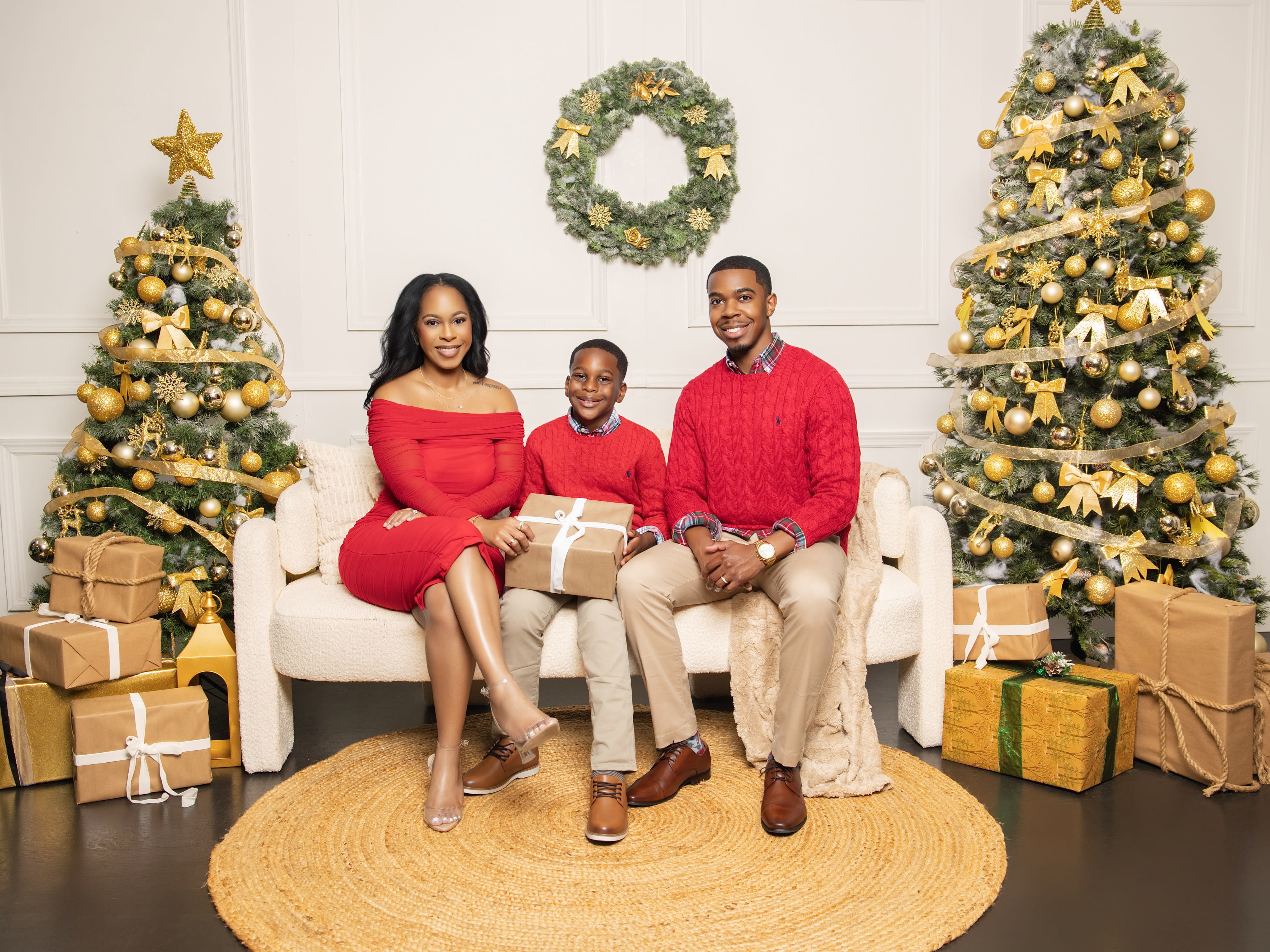 Two women in festive Christmas pajamas sit on a green couch, holding a baby, surrounded by decorated Christmas trees and holiday decorations.