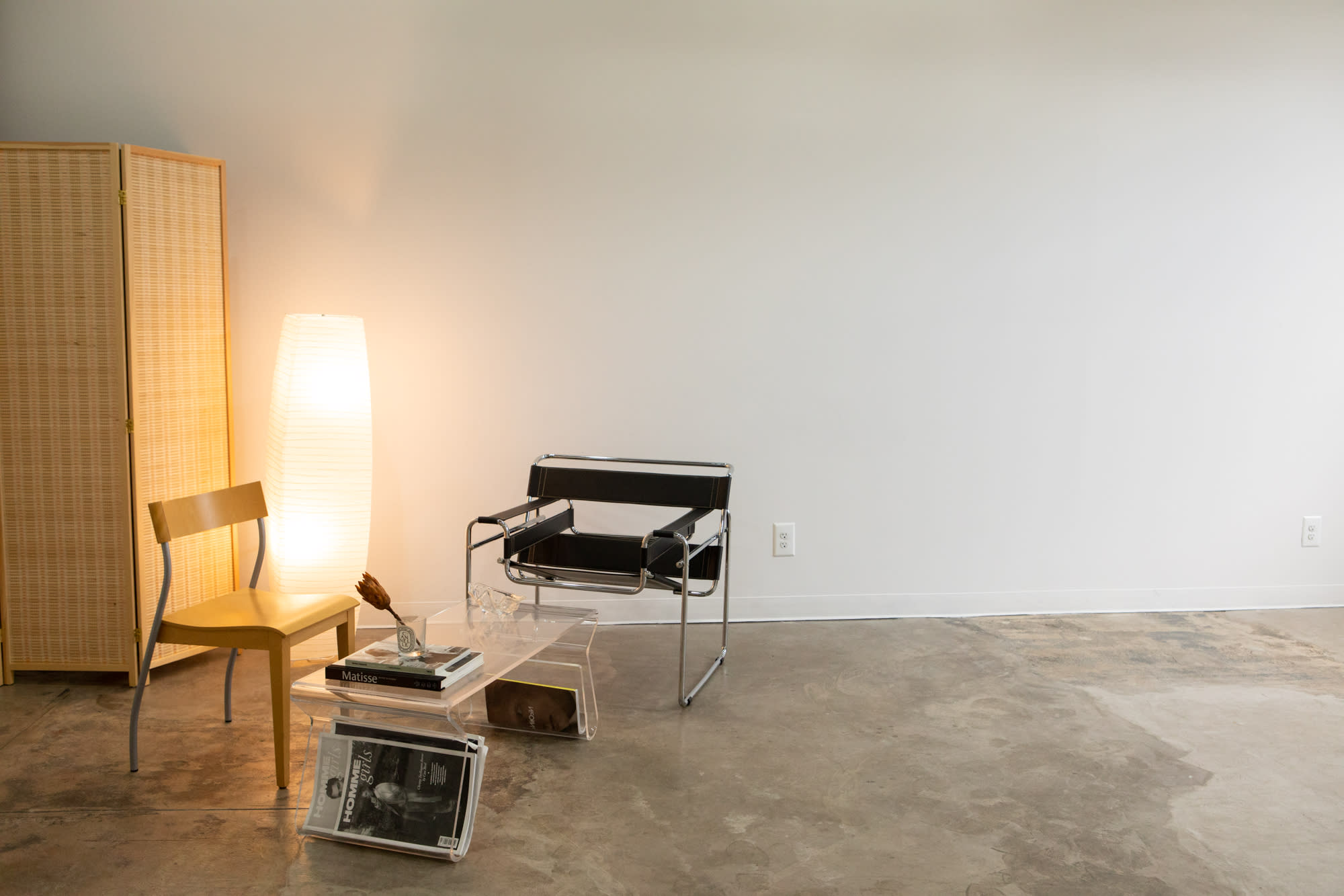A photographer captures a model seated in a chair during a photoshoot against a plain backdrop.
