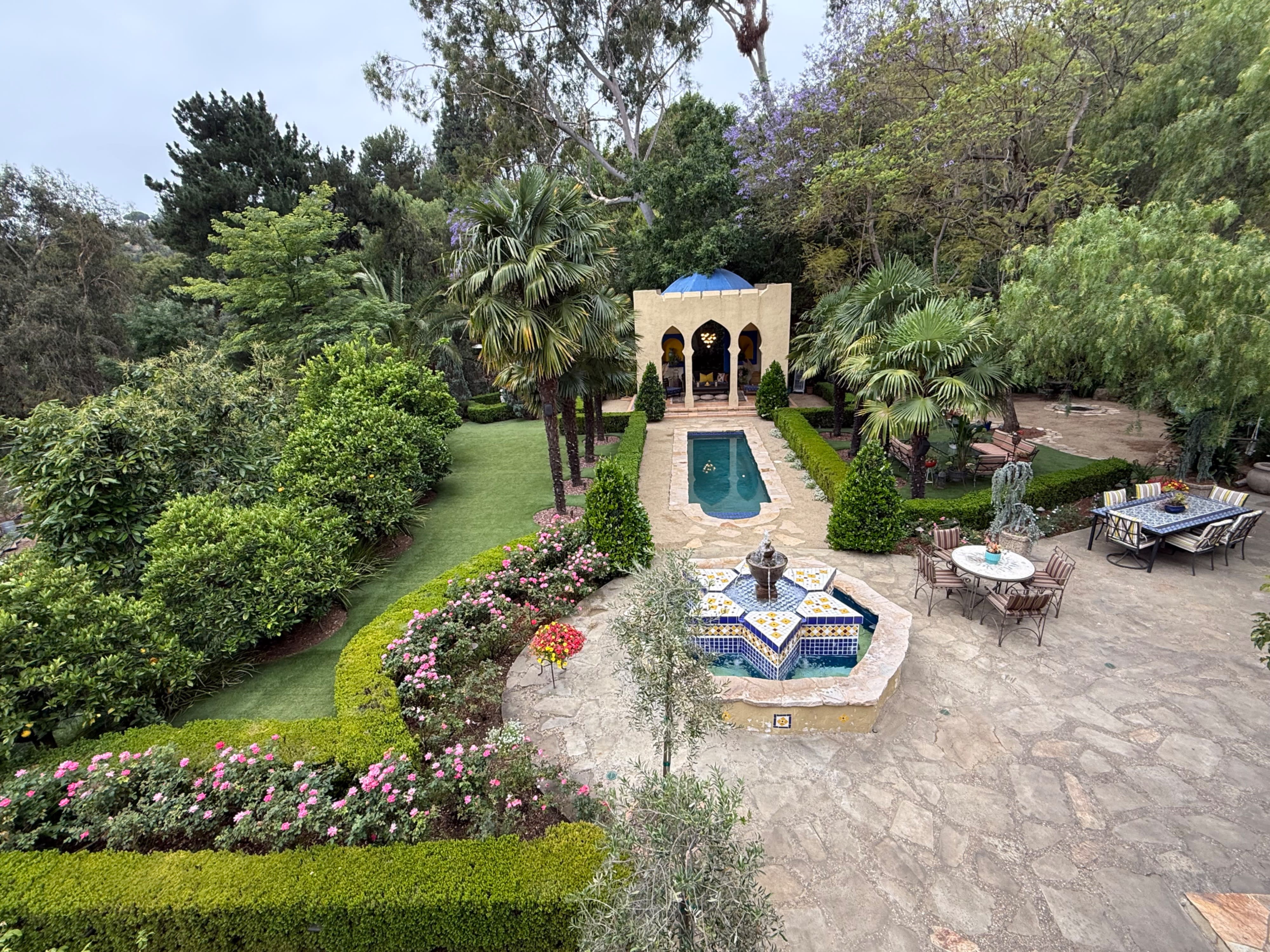 A model poses in a black outfit and wide-brimmed hat on a well-manicured lawn surrounded by greenery.