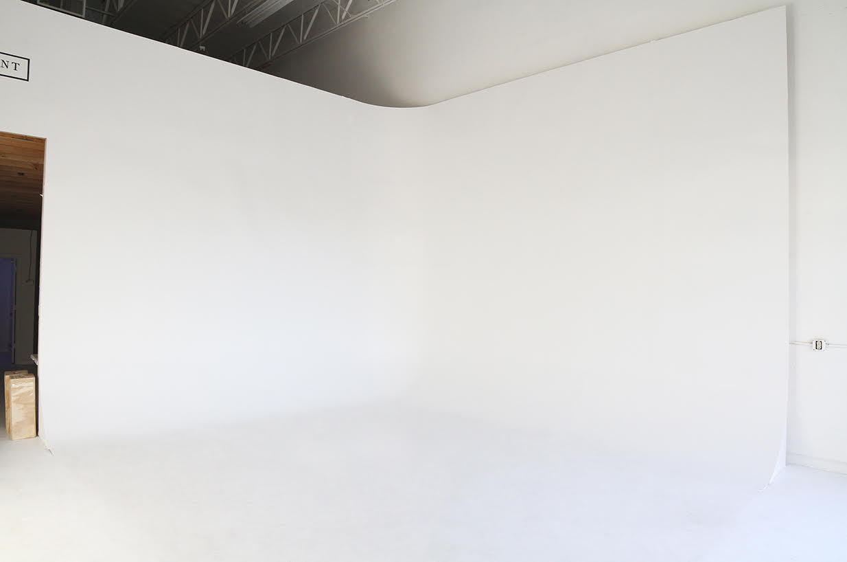A woman sits on a white pedestal, laughing while adjusting her shoe in a simple studio setting.