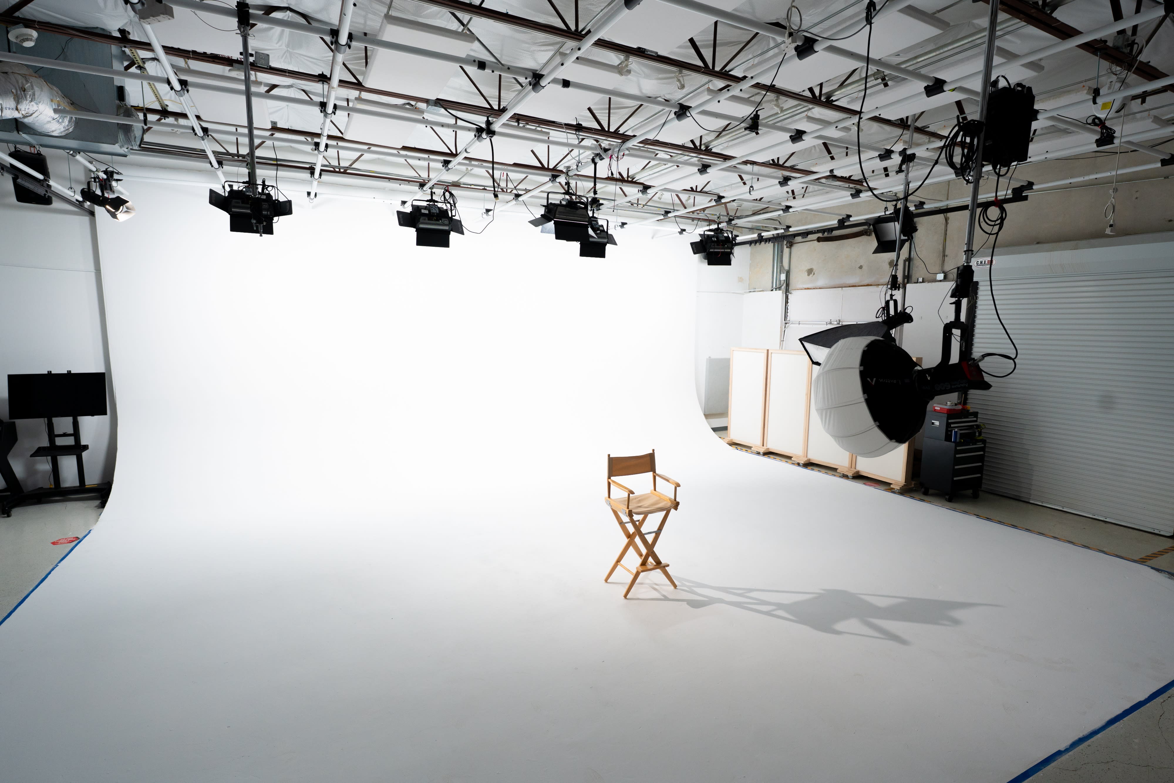 A group of seven people sits on high stools in a studio setting, engaged in a discussion with a backdrop of colorful panels.