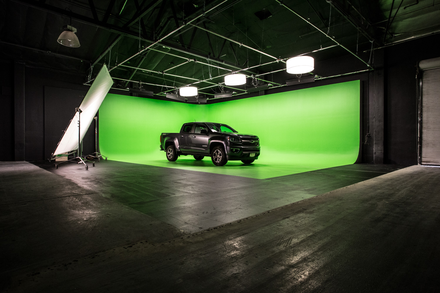 Two individuals sit inside a blue vintage car, while filming equipment and monitors are positioned in front of them against a green screen backdrop.