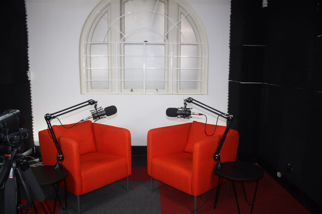 Two men sit on orange chairs in a studio setup for a podcast video shoot, with microphones positioned in front of them and a small table between them.