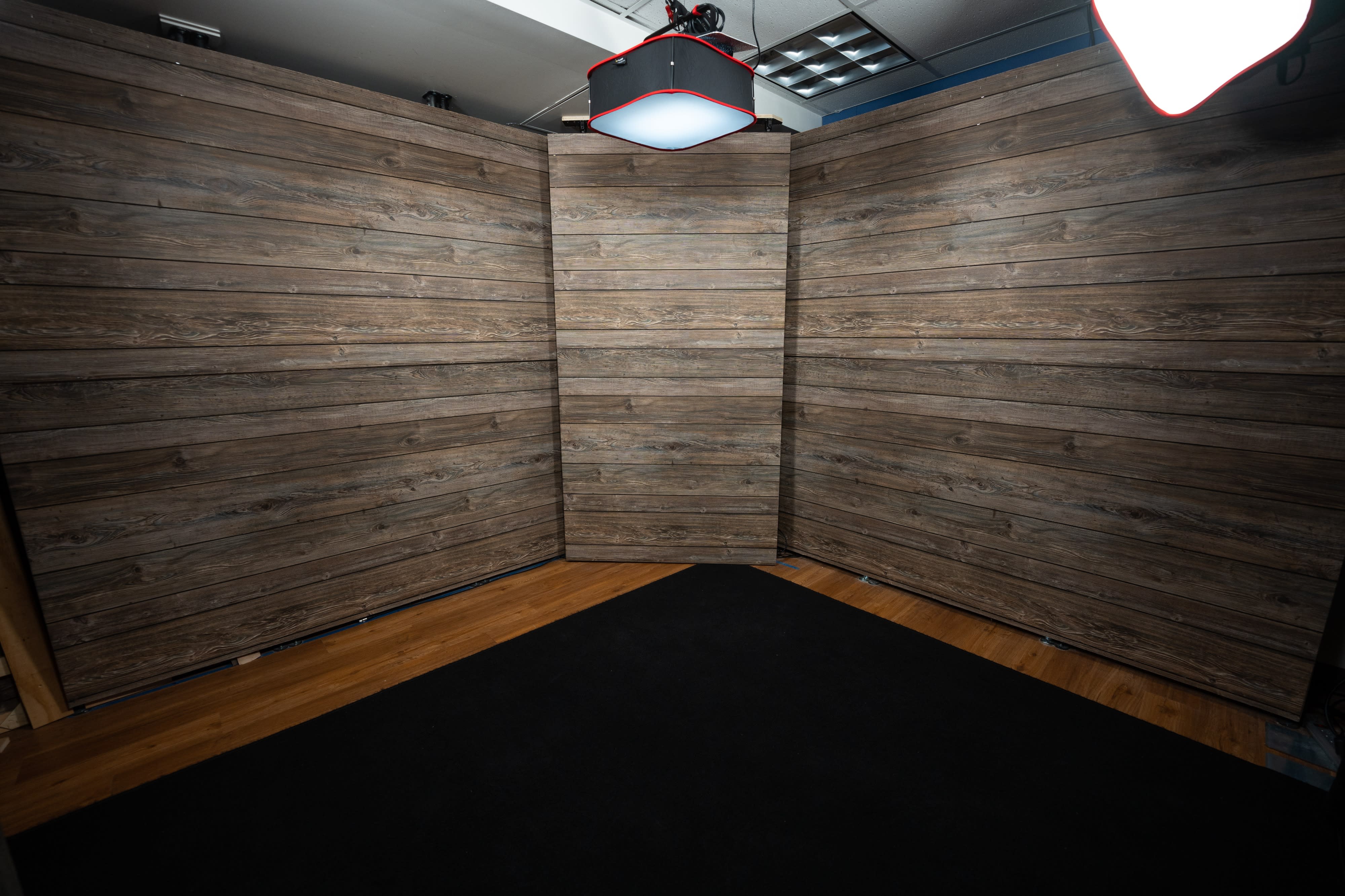 The image shows a podcast recording setup with two microphones mounted on stands positioned at a wooden table flanked by cameras, and an illuminated wooden backdrop.