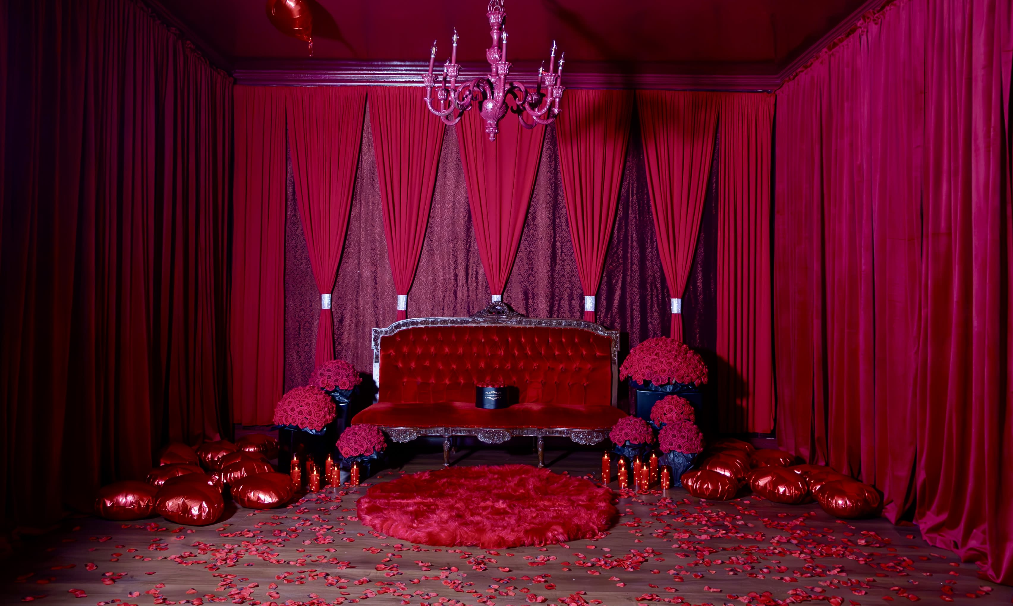 A group of fifteen women dressed in black attire poses together in a red-themed room adorned with curtains and heart-shaped decorations.