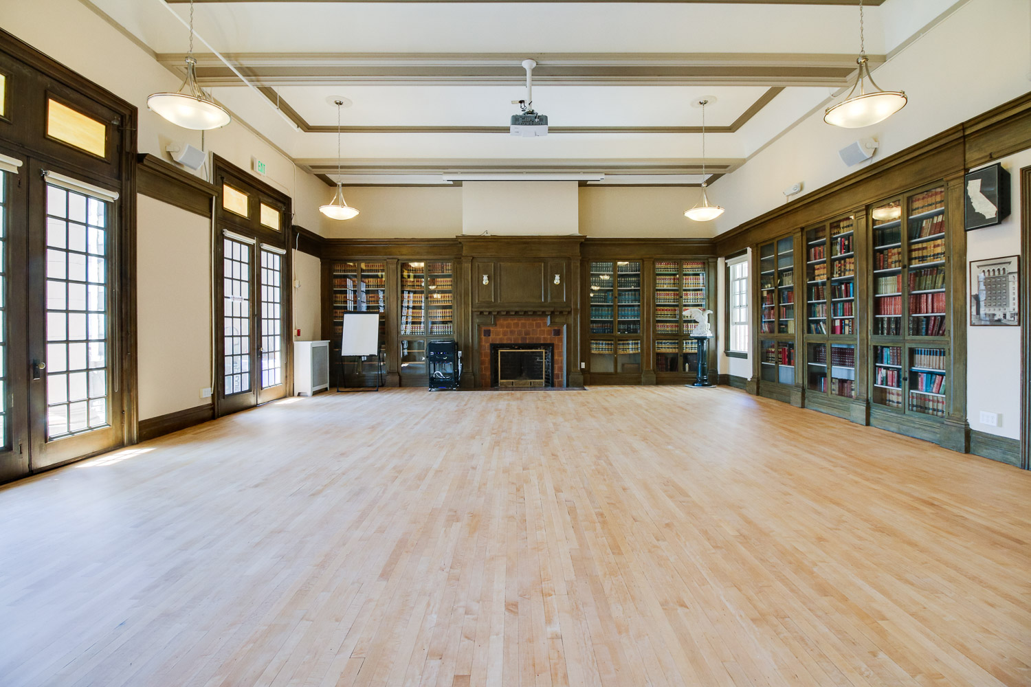 A film shoot is taking place in a room lined with bookshelves, featuring a director gesturing towards a chalkboard while a crew member adjusts equipment and another actor observes.