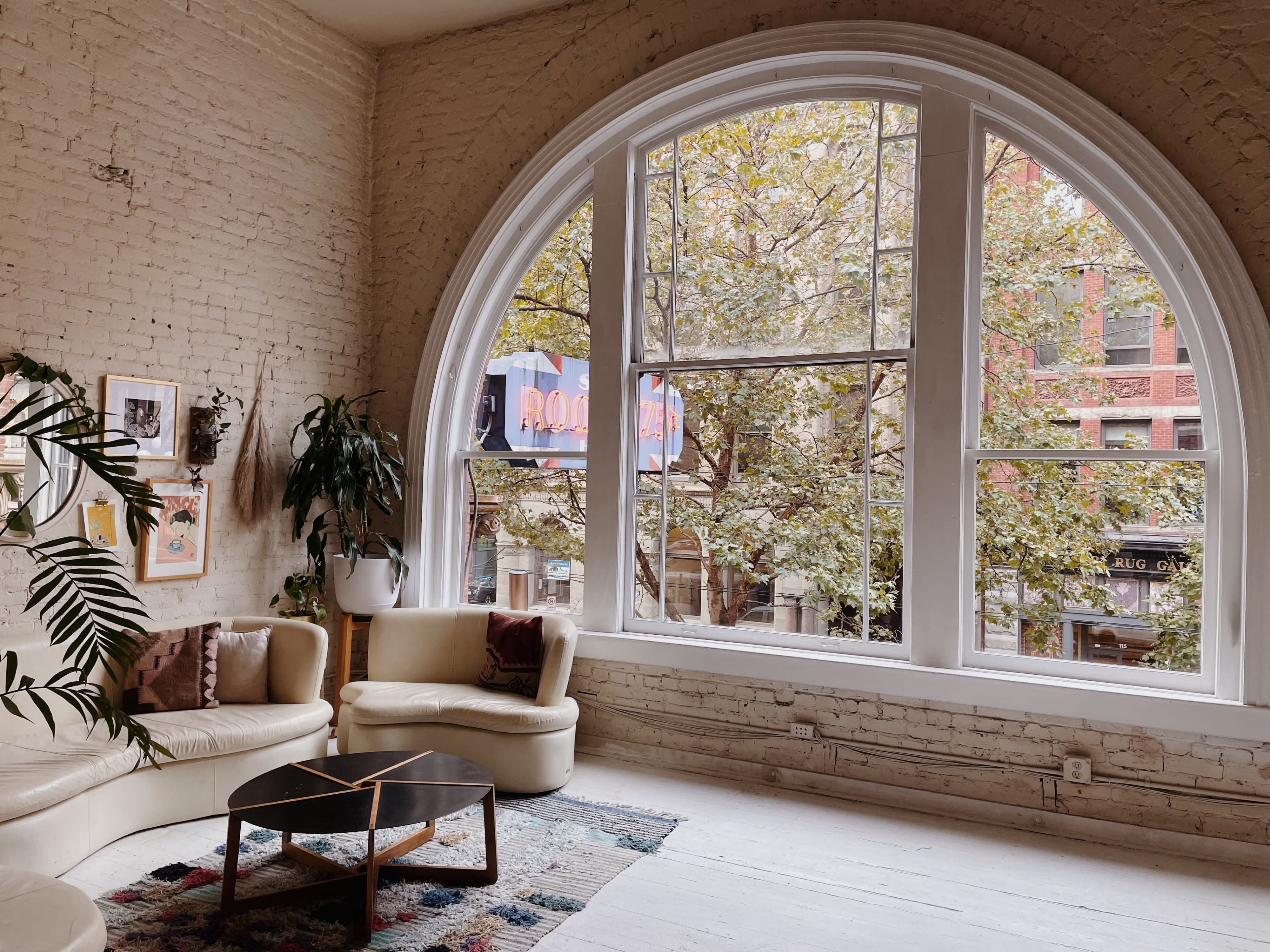 A cozy dining area features a cream-colored sectional sofa, a circular coffee table, and a large window with a view of the street, ready for a birthday dinner celebration.