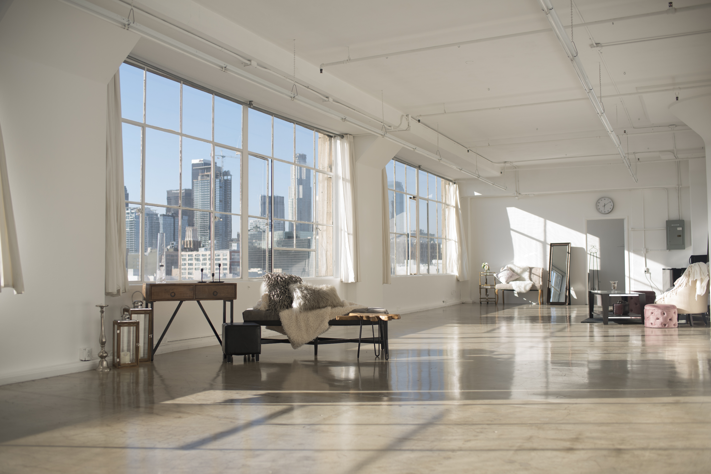 A white chaise lounge with a soft throw and pillows sits in front of large windows overlooking a city skyline.