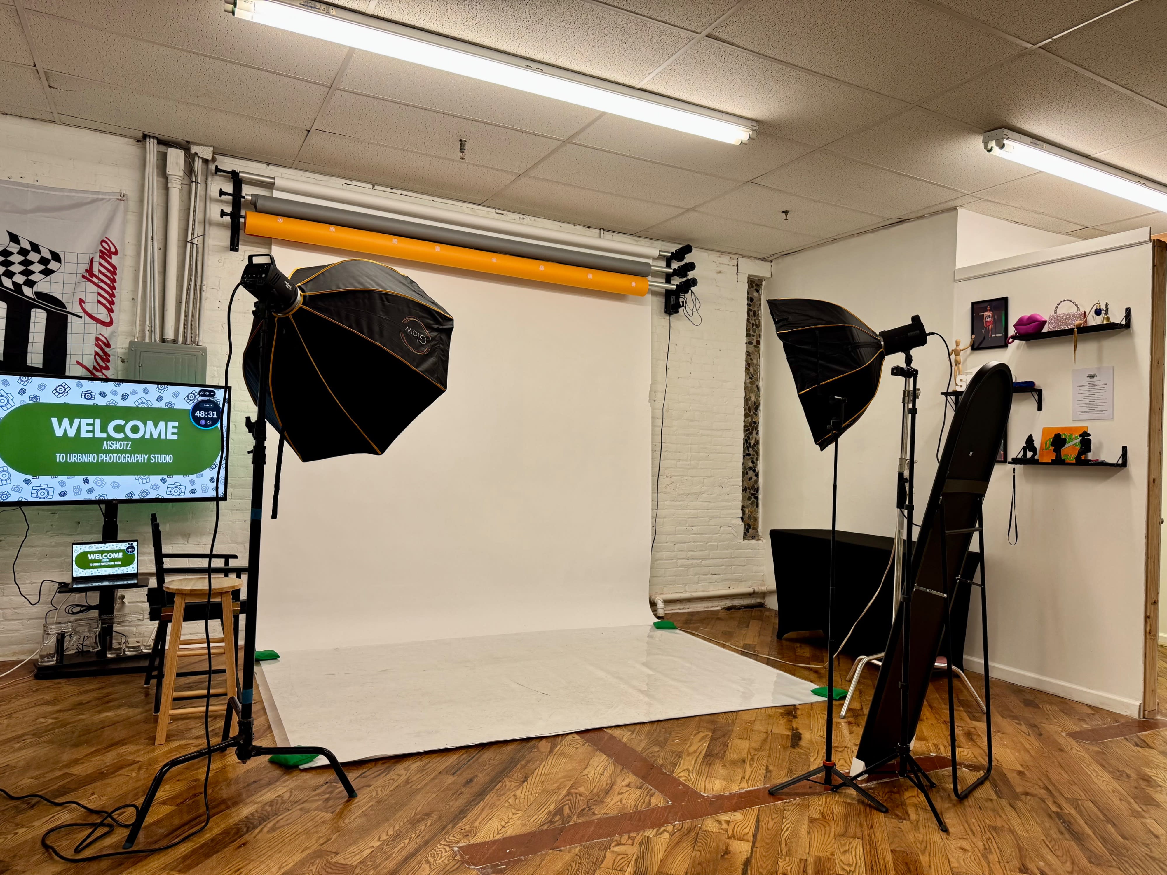 A model sits on the floor in front of a white backdrop during a photoshoot, surrounded by lighting equipment and props.
