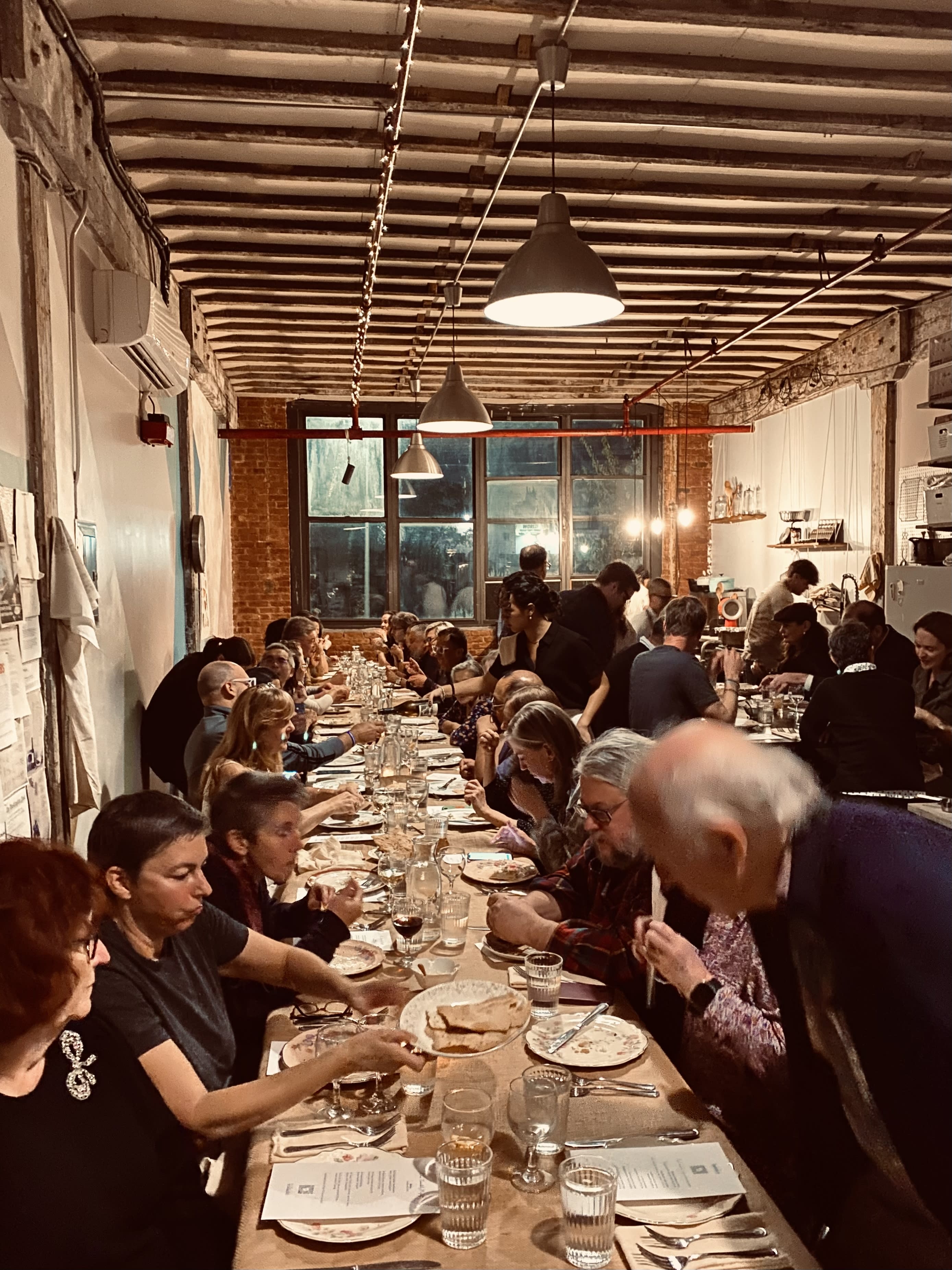 A long dining table is set with plates, utensils, and menus, illuminated by flickering candles, preparing for a dinner gathering.