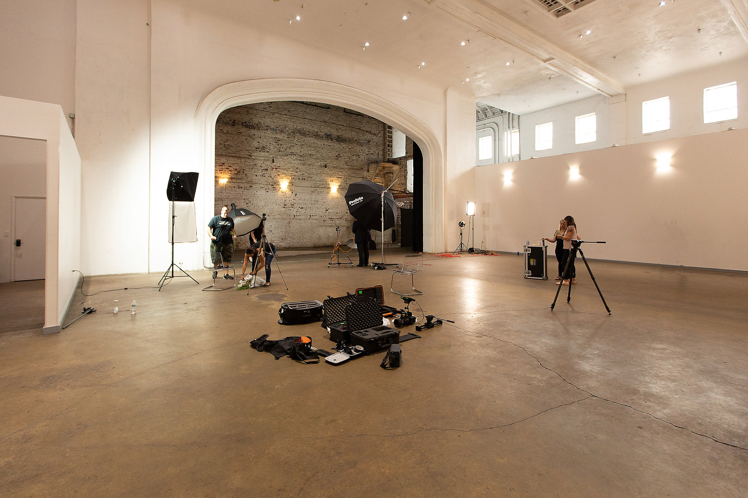 A person is filming another individual sitting on the ground in an industrial space with exposed brick walls and soft lighting.