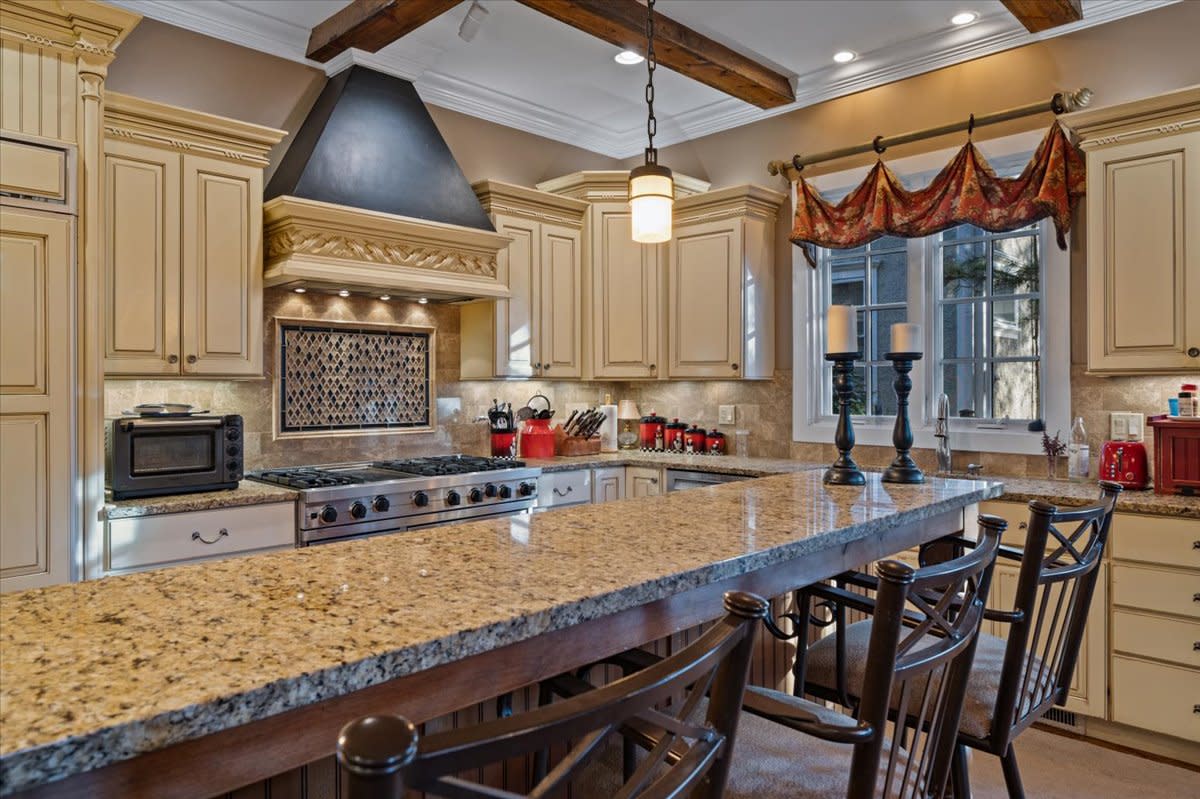 The kitchen features a wood-beamed ceiling, a central island with bar stools, and a dining table surrounded by chairs, creating a functional space for dining and gathering.