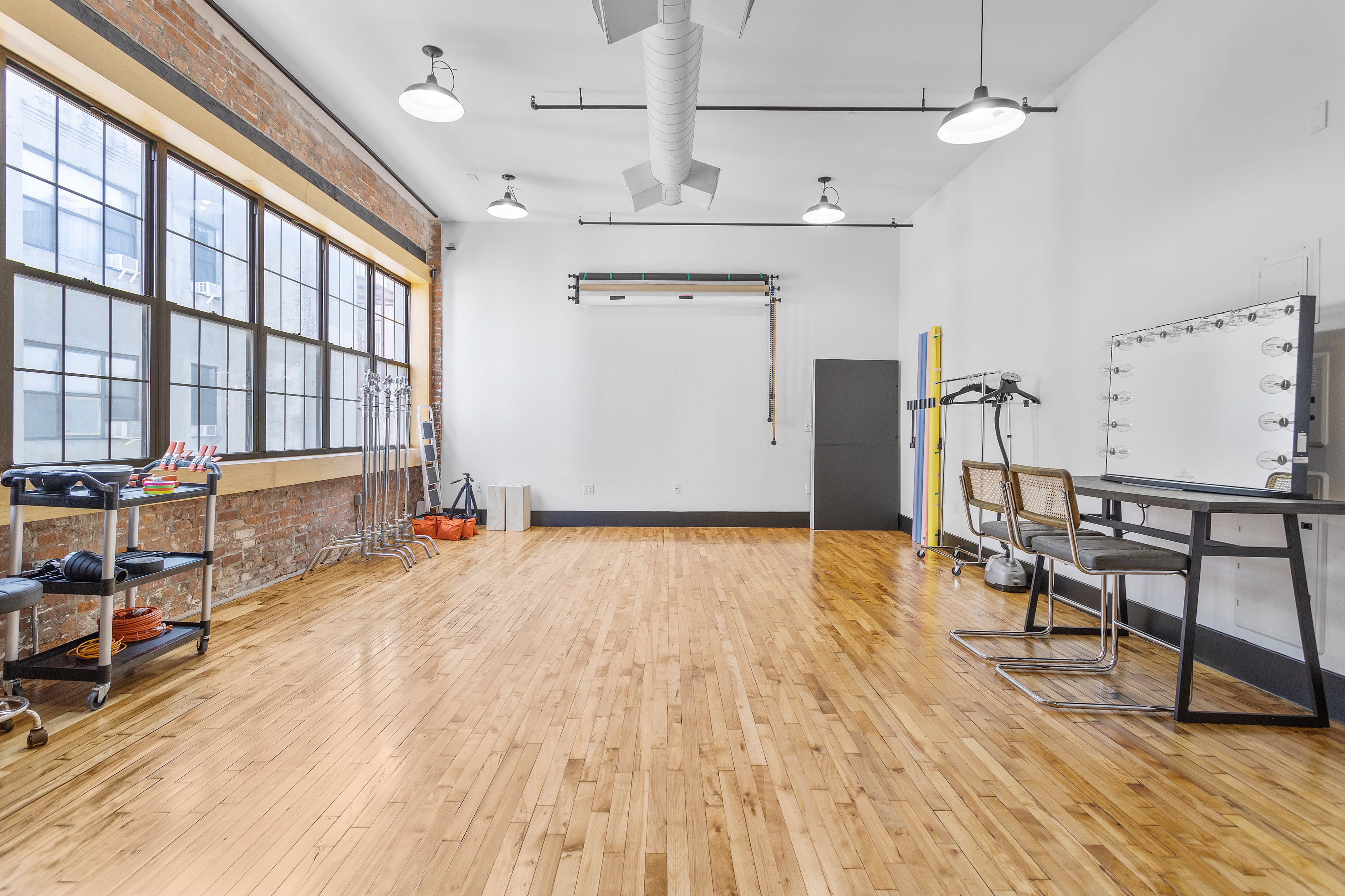 The image shows a photography studio with lighting equipment on stands, a small table with a laptop, and a backdrop setup near large windows.