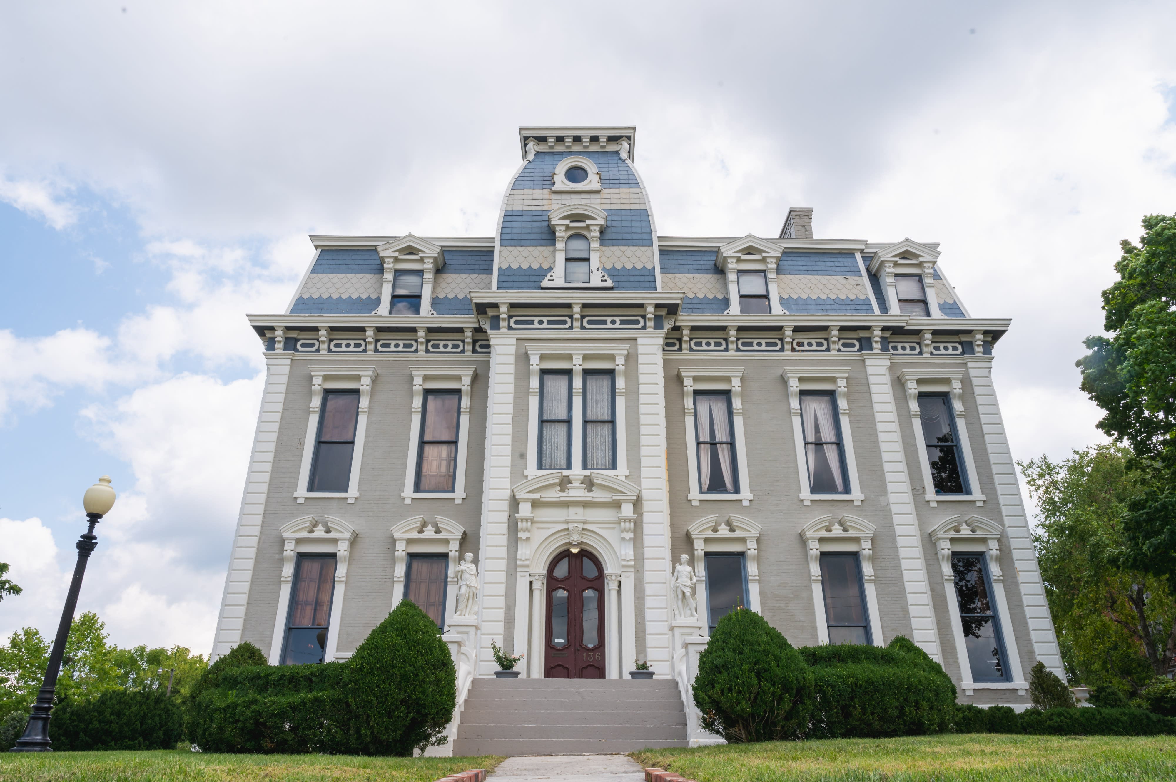 A video shoot is taking place in front of a large, elegant mansion, featuring decorative flowers and a set of chairs and a table on the lawn.