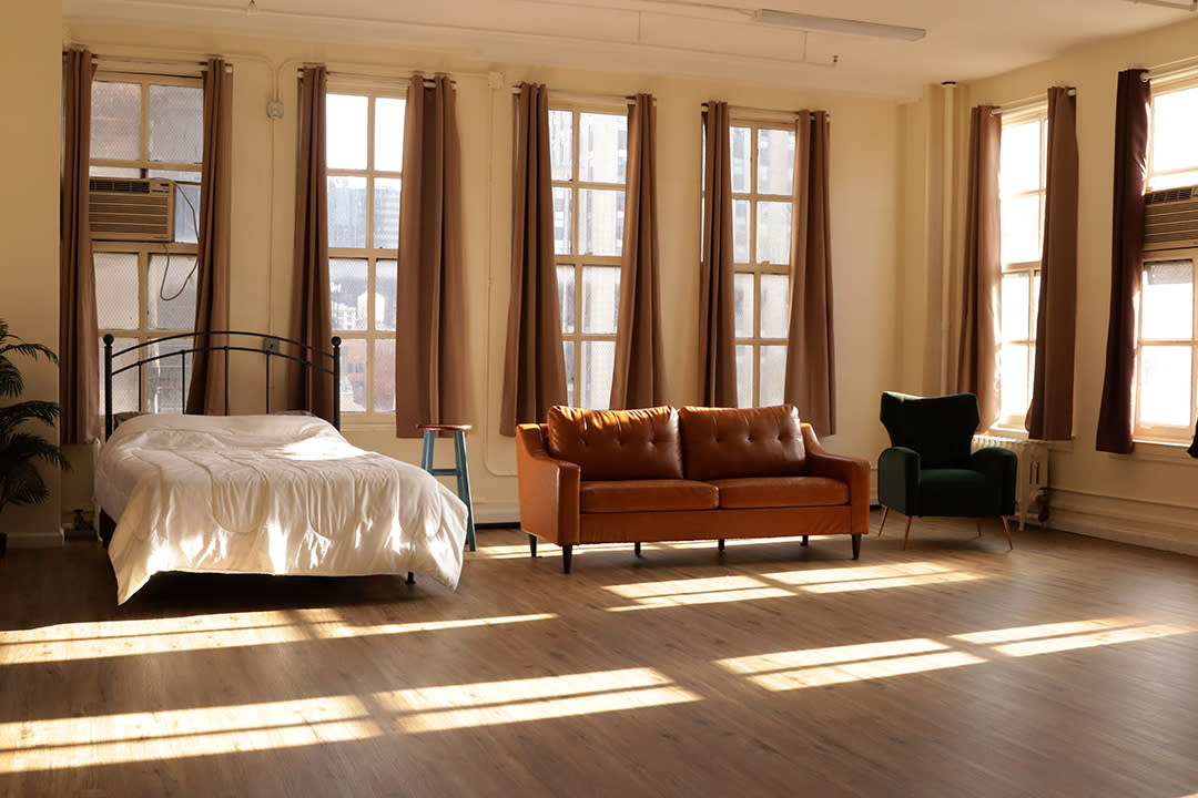 A foggy video production setup features a bed in a sparsely furnished room with curtains drawn and soft lighting aimed at the scene.