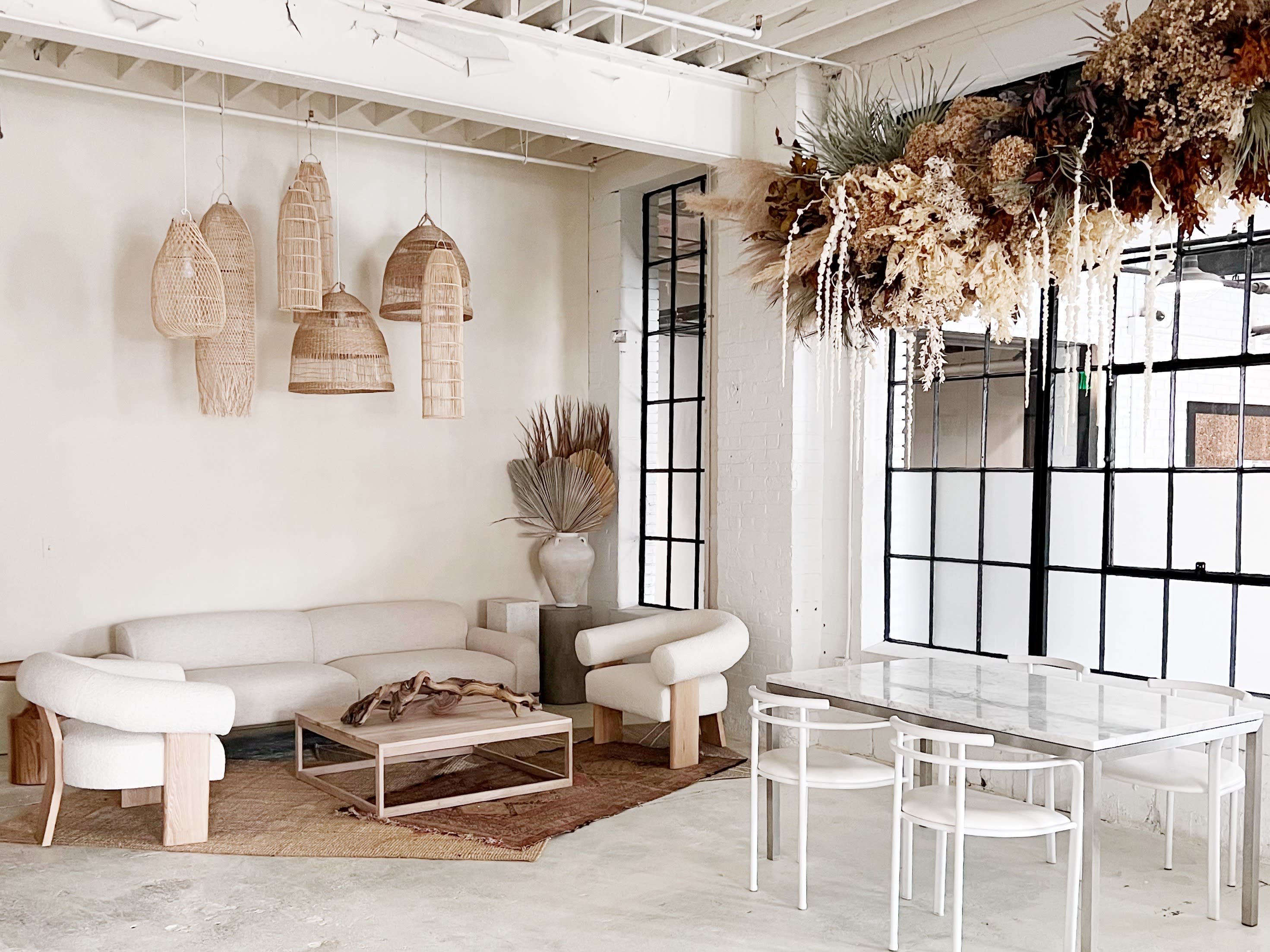 A spacious, well-lit room features a cozy seating area with a sofa and a large dining table surrounded by chairs, all under a decorative arrangement of dried plants.