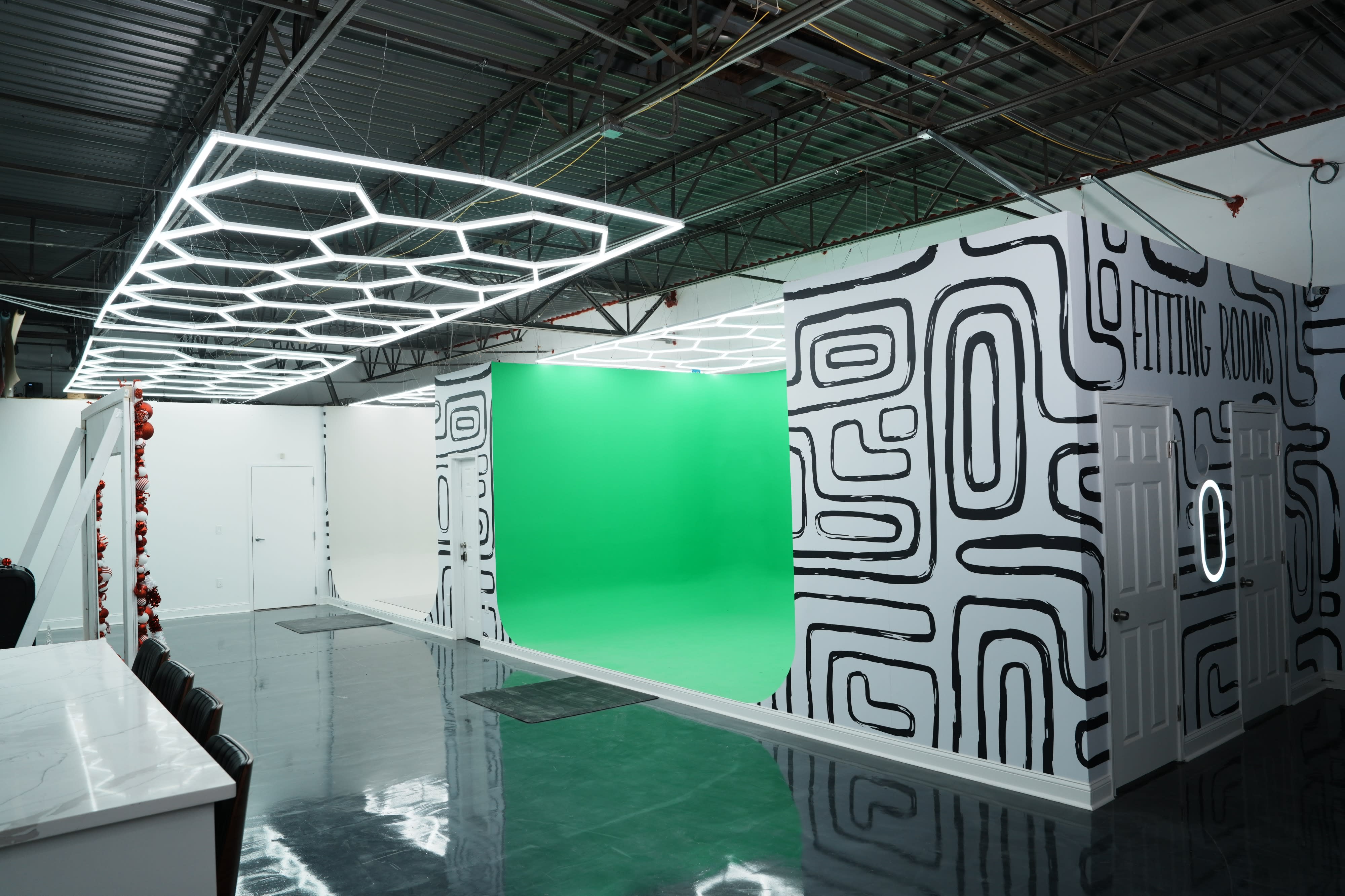 A videography shoot is set up in a studio with a bright green screen, a tripod on a small table, and a director's chair positioned nearby.