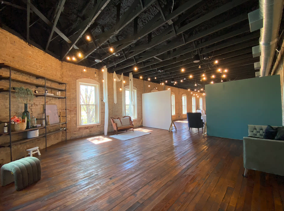 A video shoot setup is underway in a spacious, well-lit room with exposed brick walls and wooden flooring, featuring a ladder and crew members preparing equipment.