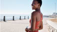 A smiling woman in athleisure standing outside near a body of water as she goes for a walk as part of her mental health routine.