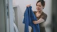 A woman holding up and folding a blue long-sleeve shirt at home in the laundry room.