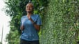 A woman calmly jogging outside near a tall green bush.