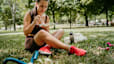 A woman eating a meal from a container while sitting in the grass outside. She's wearing workout clothes.