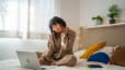 A woman experiencing tech neck. She is rubbing her neck while sitting in bed and looking down at her laptop.