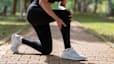 Close-up photo of a runner bent down on one knee on an outdoor path, scratching their calf and holding their knee.