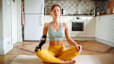 A woman meditating on her yoga mat at home in her kitchen. She's sitting in lotus position, closing her eyes, and has her hands resting on her knees.