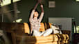 A woman stretching her arms overhead while closing her eyes and smiling. She is sitting on the couch in the sunlight.