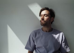 A relaxed young man leaning against a sunny white wall while closing his eyes and practicing ujjayi breathing.