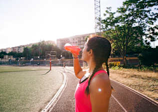 A side view of a woman drinking an orange-red electrolyte drink on an outdoor track.