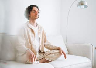 A young man sitting cross-legged on a white couch in a bright room and meditating.
