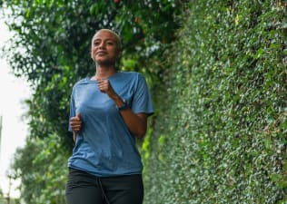 A woman calmly jogging outside near a tall green bush.