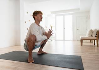 Man practicing Garland Pose (Malasana or Yoga Squat) during a Peloton yoga class at home.