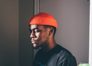 A man wearing an orange beanie sitting in the light while closing his eyes and practicing visualization meditation.