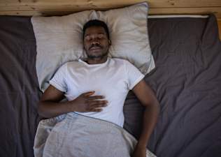 A man practicing breathing exercises for sleep. He's lying on his back in bed, eyes closed, with his right hand on his upper abdomen.