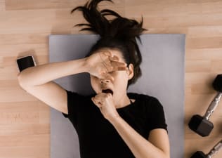 A woman yawning during a workout. She's lying down on a yoga mat next to her phone and a pair of dumbbells and covering her mouth and eyes as she yawns.