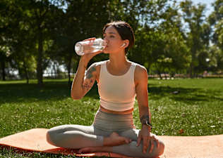 A woman with tattoos meditating and drinking water while sitting on a yoga mat outdoors.