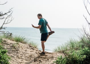 A man watching a video on his phone with wire headphones while simultaneously doing a standing quad stretch. He is standing on a beautiful sandy trail near the ocean.