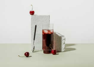 A glass of tart cherry juice propped against a cinder block with tart cherries and a spoon nearby.