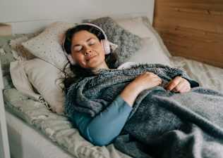 A relaxed woman meditating in bed. She has headphones on and is under a blanket.