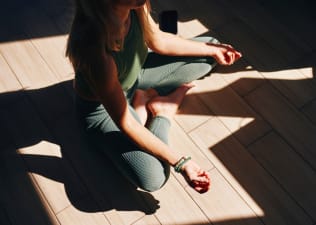 A close-up photo of a woman practicing mantra meditation at home. She's sitting in a criss-cross position with her hands and arms resting on her legs.