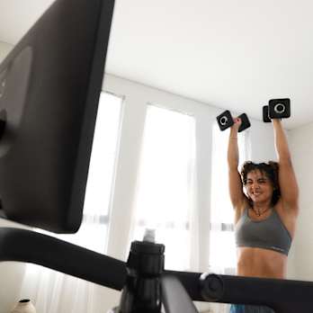 woman doing a shoulder press with dumbbells next to a Peloton Tread